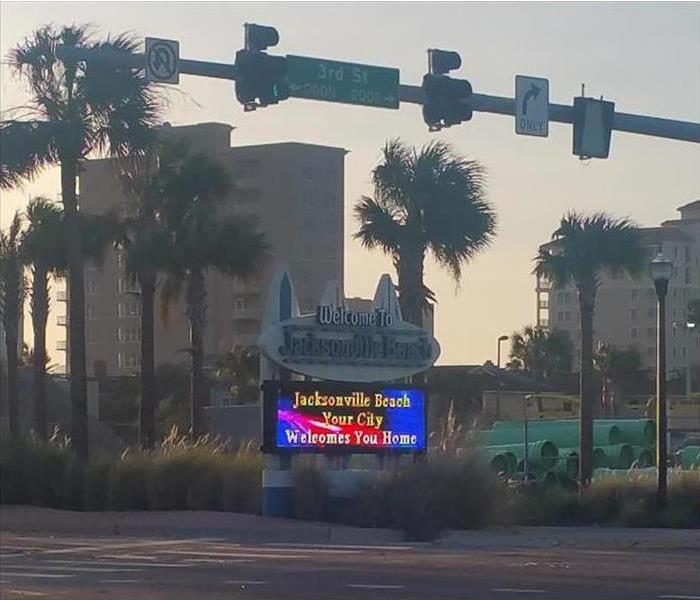 Image shows a lighted sign in an intersection with a traffic light and street sign over head and a palm tree and two condos.
